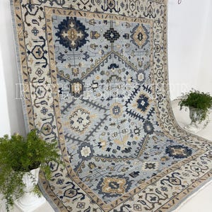 Puede incluir: Una alfombra rectangular grande con un campo central azul claro y patrones geométricos intrincados en azul marino, beige y dorado. La alfombra tiene un borde detallado y flecos a lo largo de los bordes, creando un aspecto clásico y elegante.