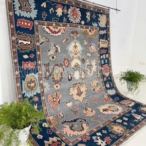 May include: A large, rectangular rug with a central gray field and a dark blue border. The rug features intricate floral and geometric patterns in shades of pink, beige, and brown. The rug is displayed against a white background.