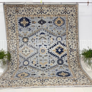Puede incluir: Una alfombra rectangular grande con un campo central azul claro y patrones geométricos intrincados en azul marino, beige y gris. La alfombra presenta un borde detallado y se exhibe sobre una superficie blanca. El texto "RUGGERS STORE" es visible.