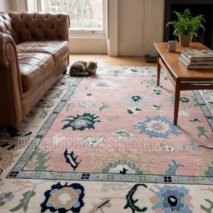 May include: A pink area rug with a floral pattern in blue, green, and cream. The rug has a border with a repeating design and the text "RUGUERESTORE" across the center. The rug is in a living room setting.