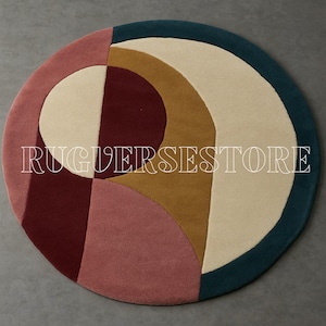 Puede incluir: Una alfombra redonda con un diseño geométrico. La alfombra presenta un borde verde azulado, con secciones crema, beige, burdeos y rosa. El texto "RUGERSSTORE" se muestra en blanco en el centro de la alfombra.