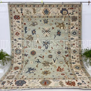 May include: A large, rectangular rug with a pale blue central field and a detailed border. The rug features intricate patterns in beige, brown, red, and blue. The text "RUGERSSTORE" is displayed across the center of the rug.