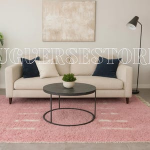 May include: A living room scene featuring a light-colored sofa with navy and cream-colored pillows, a round black coffee table, and a pink rug with white accents. A black floor lamp and a potted plant are also visible.