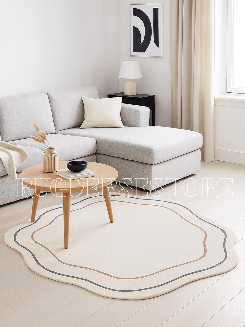 May include: A cream-coloured, wavy-edged rug with brown and grey border lines. The rug is in a living room with a light-coloured sofa, wooden coffee table, and vase. The room has a minimalist design. The rug's shape is irregular.