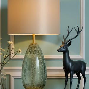 Peut inclure: Une lampe de table décorative avec un abat-jour beige et un pied en verre vert texturé. Une figurine de cerf noire se trouve à proximité. La lampe a une tige et une base dorées, sur un mur vert clair.