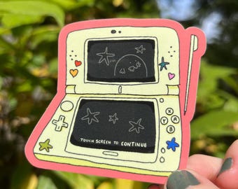 May include: A pale yellow handheld gaming console sticker with a pink border. The top screen shows a star and a creature, while the bottom screen displays the text TOUCH SCREEN TO CONTINUE. A stylus is attached to the right side of the device.