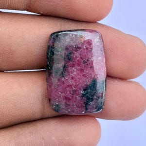 May include: A rectangular cabochon gemstone featuring a mottled pattern of deep magenta, dark green, and black mineral inclusions. The stone has a smooth, polished surface with a glossy finish, displaying a natural crystalline texture throughout.