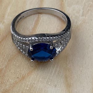 May include: A silver ring with a large, oval, dark blue gemstone as the centerpiece. The band is detailed with small, clear stones. The ring is set on a light-colored wooden surface. The brand name "Sam & Co" is visible on the ring.