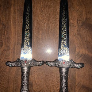 May include: Two decorative daggers with ornate silver handles and black blades. The blades feature gold and teal filigree designs. Each dagger has a red gem in the center of the handle guard and teal gems on either side.