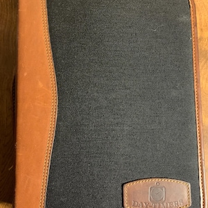 May include: A black and brown Day-Timers organizer. The organizer has a black fabric center with brown leather accents and trim. The Day-Timers logo is on a brown leather patch.