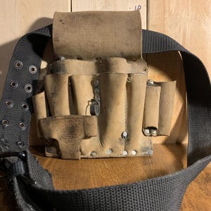 May include: A well-worn, tan leather tool belt with multiple pockets and a flap closure. The belt has a black nylon strap with metal grommets and a buckle. The tool belt is designed for carrying various tools.