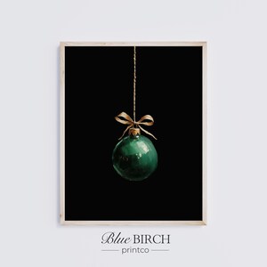 May include: A framed print featuring a single, glossy, emerald green Christmas ornament. The ornament hangs from a thin, gold-colored string and is adorned with a gold bow. The background is solid black, and the text "Blue BIRCH printco" is at the bottom.