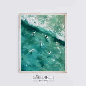 May include: Framed art print featuring an aerial view of surfers in turquoise ocean water. The artwork depicts several surfers on white surfboards near breaking waves. The print is framed in a light wood frame. The text "BlueBIRCH printco" is at the bottom.