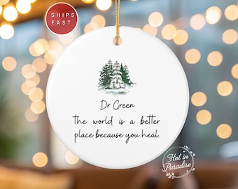 May include: White ceramic ornament with a watercolor image of evergreen trees and the text "Dr Green the world is a better place because you heal." A gold string is attached for hanging.