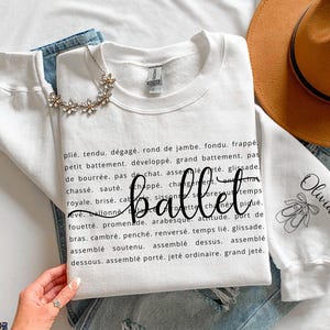 May include: A white crewneck sweatshirt featuring a list of black printed ballet terms, the word ballet in a large cursive script, and a sleeve graphic of ballet slippers with the name Olivia printed in black ink on the fabric.