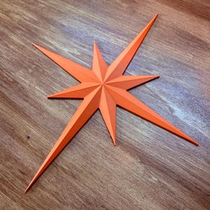 May include: An orange starburst decoration with eight long, pointed rays. The star is made of folded paper or cardstock and is set against a wooden background. The star's color is a vibrant, solid orange.