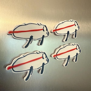 May include: Four white, cartoonish buffalo-shaped magnets with a red stripe across the body and dark blue outlines. The magnets have dark blue legs and are arranged on a silver surface.