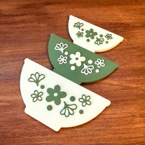 May include: Three decorative, semi-circular coasters with floral designs. Two coasters are white with green floral patterns, and one is green with white floral patterns. The coasters are arranged on a wooden surface.