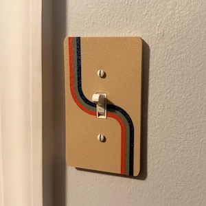 May include: Tan light switch cover with a retro design. The cover features a curved stripe design in red, green, and navy blue. The switch is in the "on" position.