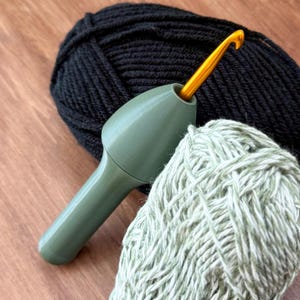 May include: A green crochet hook with a gold hook tip is inserted into a green handle. Two balls of yarn are in the background, one black and one light green.