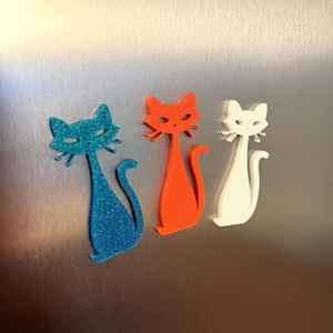 May include: Three stylized cat figurines in blue glitter, orange, and white are displayed. The cats have long, slender bodies and are posed with their tails curled. The background is a brushed metal surface.