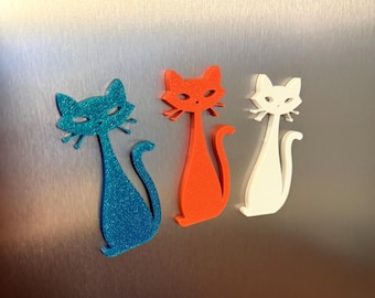 May include: Three stylized cat figurines in blue glitter, orange, and white are displayed. The cats have long, slender bodies and are posed with their tails curled. The background is a brushed metal surface.