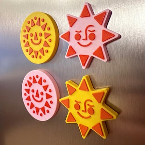 May include: Four colorful sun-shaped magnets. Two are yellow with orange accents, and two are pink with orange accents. Each sun has a smiling face and triangular rays. These decorative magnets are perfect for the kitchen or office.