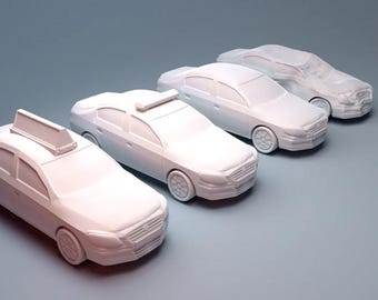 May include: Four white toy cars are arranged diagonally on a gray surface. The cars include a taxi and three sedans. The taxi has a roof sign. The cars have detailed features like wheels, headlights, and grilles.