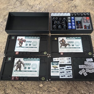 Könnte beinhalten: Ein schwarzer Spiel-Organizer mit mehreren Fächern, darunter eine Aufbewahrungsbox, Würfelhalter und Tabletts für Karten und Spielsteine. Die Tabletts enthalten Spielkarten mit Text und Illustrationen von Charakteren. Der Organizer ist für Tabletop-Gaming konzipiert.