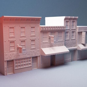 Puede incluir: Maqueta beige de una manzana de edificios, con tres edificios con detalles de ladrillo, ventanas y toldos. El primer edificio tiene el texto "Murphy's" en la fachada. El modelo representa un distrito comercial de un pueblo pequeño.