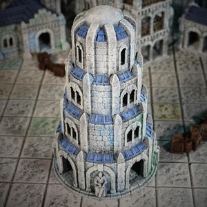 Peut inclure: Une tour miniature détaillée, conçue pour les jeux de société. La tour présente plusieurs niveaux, des fenêtres cintrées et des détails bleus. Elle est faite pour ressembler à de la pierre, avec un toit en dôme. D'autres bâtiments sont visibles en arrière-plan.