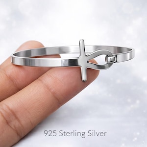 Puede incluir: Un brazalete plateado con un símbolo Ankh. El brazalete está hecho de metal pulido y tiene un diseño sencillo y elegante. El texto "925 Sterling Silver" es visible en la parte inferior de la imagen. Una joya elegante.