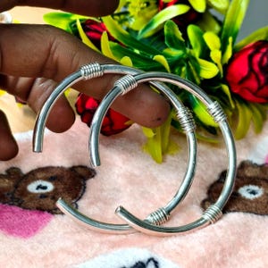 Puede incluir: Dos brazaletes plateados de diseño abierto. Cada brazalete presenta un diseño de alambre enrollado en el centro. El fondo incluye una mano, flores y una superficie rosa con un patrón de oso.