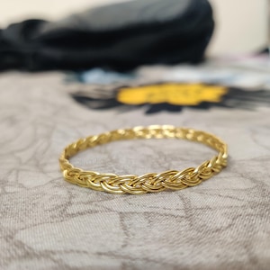 Può includere: Un braccialetto intrecciato color oro. Il braccialetto ha un design intrecciato ed è circolare. Il gioiello è su una superficie a fantasia. Il testo "treeSilver925" è visibile sul braccialetto.