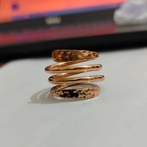 May include: A copper-colored spiral ring with a hammered texture. The ring wraps around the finger in a continuous coil, with a slightly irregular surface. The ring is made of a shiny metal.