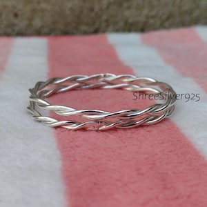 May include: A silver-colored ring with a braided design. The ring is made of shiny metal and features two intertwined strands. The ring is displayed on a pink and white striped surface. The text "ShreeSilver925" is visible.
