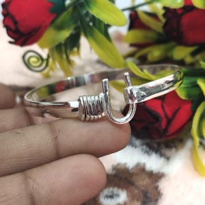Op de afbeelding: Een zilverkleurige armband met een unieke haak- en oogsluiting. De armband heeft een gewikkeld draadontwerp bij de sluiting. De achtergrond bevat rode rozen en groen blad.