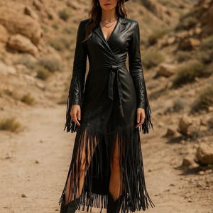 May include: A black leather wrap dress featuring long sleeves with fringe detailing, a tie waist, and a high-low hemline adorned with long black fringe. The garment is paired with a black wide-brimmed hat and black boots, creating a cohesive look.