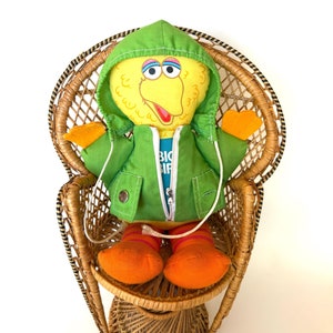 May include: A yellow Big Bird plush toy wearing a green hooded jacket with a zipper and a blue shirt that says "Big Bird" is sitting in a wicker chair.