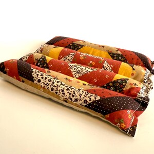 May include: A rectangular, quilted pillow with a patchwork design. The pillow features a variety of fabrics in shades of red, yellow, brown, and cream, with floral and polka dot patterns. The pillow is approximately 30 cm long and 20 cm wide.