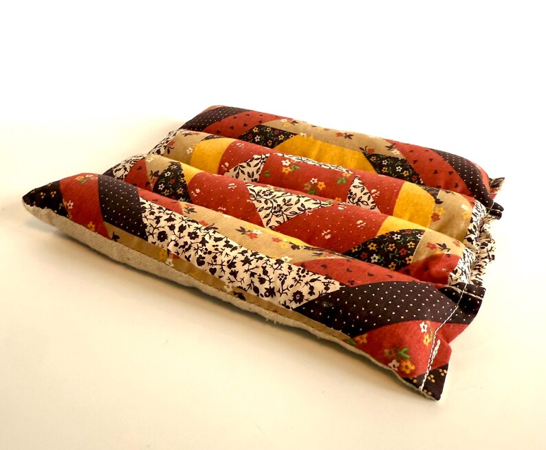 May include: A rectangular, quilted pillow with a patchwork design. The pillow features a variety of fabrics in shades of red, yellow, brown, and cream, with floral and polka dot patterns. The pillow is approximately 30 cm long and 20 cm wide.