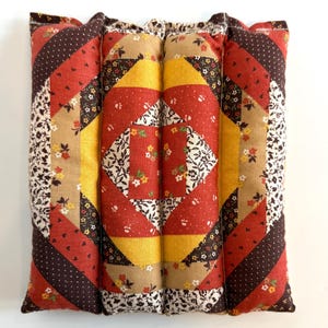 May include: A rectangular, quilted pillow with a patchwork design. The pillow features a combination of red, yellow, brown, and floral patterned fabrics. The design includes diagonal stripes and square patterns, creating a vintage aesthetic.