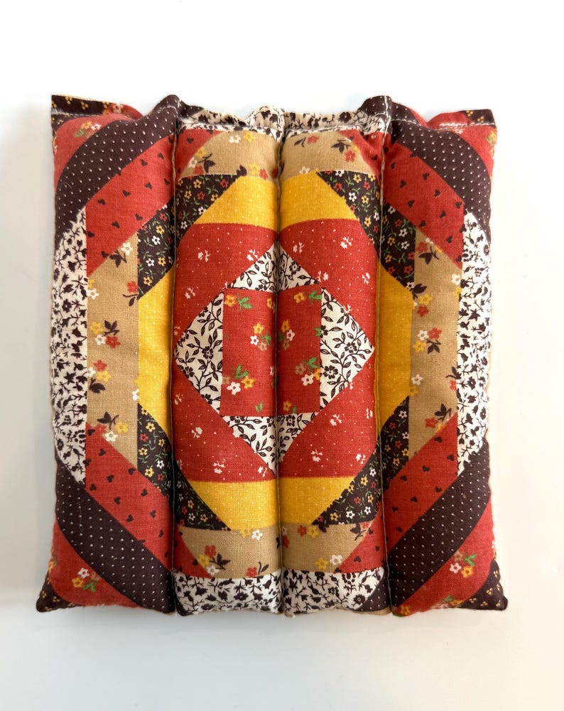 May include: A rectangular, quilted cushion with a patchwork design. The cushion features red, yellow, brown, and floral patterned fabrics. The design includes diagonal stripes and square patterns, creating a vintage aesthetic.