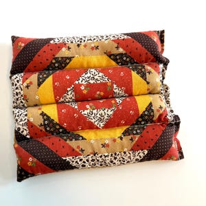 May include: Four stacked rectangular pillows with a patchwork design. The pillows feature red, yellow, brown, and floral patterned fabrics. The design includes diagonal stripes and square patterns, creating a vintage aesthetic.