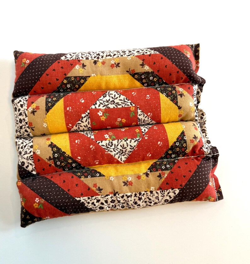 May include: Four stacked rectangular pillows with a patchwork design. The pillows feature red, yellow, brown, and floral patterned fabrics. The design includes diagonal stripes and square patterns, creating a vintage aesthetic.