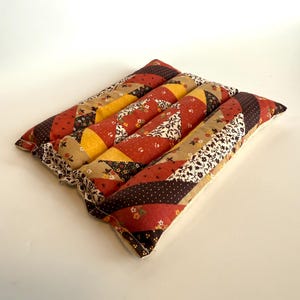 May include: A patchwork cushion with a square shape. It is made from various fabrics in red, brown, yellow, and beige, with floral and polka dot patterns. The edges are finished with a ruffled trim. The cushion is approximately 30 cm square.