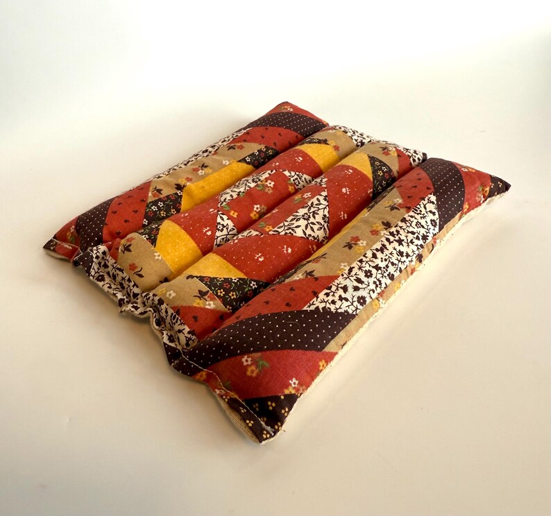 May include: A patchwork cushion with a square shape. It is made from various fabrics in red, brown, yellow, and beige, with floral and polka dot patterns. The edges are finished with a ruffled trim. The cushion is approximately 30 cm square.