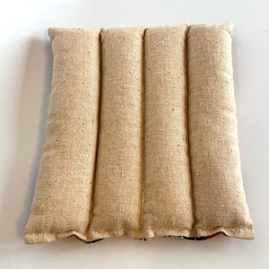 May include: A beige rectangular heating pad with four vertical, padded sections. The fabric has a textured, woven appearance. The bottom edge reveals a patterned fabric with red and blue accents. Designed for therapeutic use, it is approximately 25 cm wide.