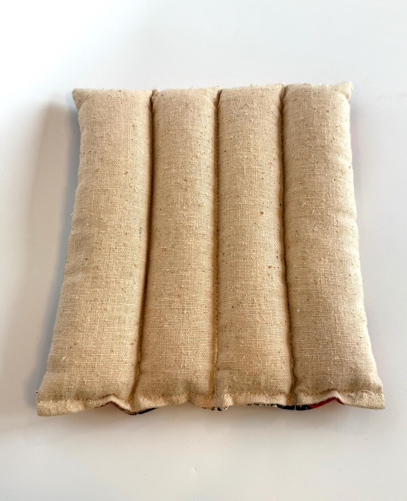 May include: A beige rectangular heating pad with four vertical, padded sections. The fabric has a textured, woven appearance. The bottom edge reveals a patterned fabric with red and blue accents. Designed for therapeutic use, it is approximately 25 cm wide.