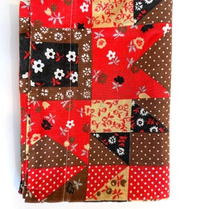 May include: Folded patchwork fabric with a vibrant mix of patterns. The design features squares and triangles in red, brown, black, and tan, adorned with floral and polka dot motifs. The fabric is folded neatly, showcasing the variety of patterns.